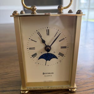 Op de afbeelding: Een rechthoekige quartzklok van goudkleurig metaal met een handvat aan de bovenkant. De gebroken witte wijzerplaat heeft zwarte Romeinse cijfers, zwarte wijzers en een blauwe maanfase-indicator met gouden sterren.