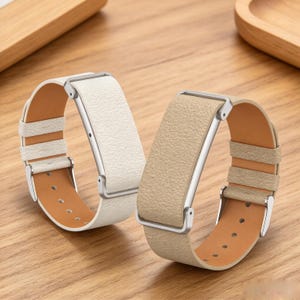 Op de afbeelding: Twee horlogebandjes met een textuur, één in wit en één in beige. Elk bandje heeft een zilverkleurige metalen connector en een bijpassende zilverkleurige gesp. De binnenkant van beide bandjes is van glad, lichtbruin materiaal met gaatjes.