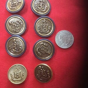 May include: Nine gold-colored buttons with a black background and a crest design. The buttons have a gold rim and are arranged in a semi-circle on a red surface.
