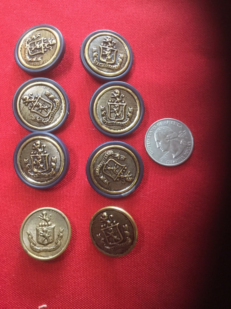 May include: Nine gold-colored buttons with a black background and a crest design. The buttons have a gold rim and are arranged in a semi-circle on a red surface.