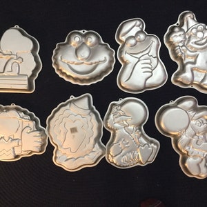 May include: Set of nine silver-colored metal cake pans featuring Sesame Street characters. The pans are shaped like Cookie Monster, Big Bird, Elmo, Bert, Ernie, Oscar the Grouch, Grover, and two other characters.