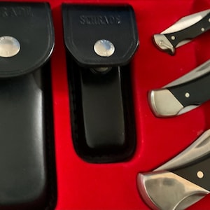 May include: A set of Schrade knives and sheaths in a red box. The knives have black handles and silver blades. The sheaths are black leather with silver snap closures. The word "Schrade" is printed on the sheaths.
