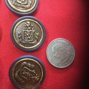May include: Four gold-colored buttons with a blue rim and a crest design. The buttons are on a red surface with a silver quarter for scale.