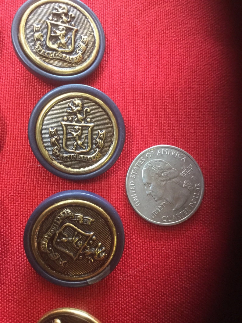 May include: Four gold-colored buttons with a blue rim and a crest design. The buttons are on a red surface with a silver quarter for scale.