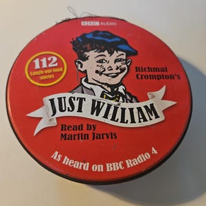 May include: A round red metal tin featuring a graphic of a boy in a blue cap. The text reads BBC AUDIO, 112 Laugh-out-loud stories, Richmal Crompton's JUST WILLIAM, Read by Martin Jarvis, and As heard on BBC Radio 4. The surface shows signs of wear.