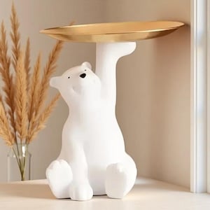 Peut inclure: Une figurine d'ours blanc en position assise avec un bras levé vers le haut pour soutenir un plateau circulaire en métal doré lisse. L'ours a une finition mate, un petit nez noir et deux yeux noirs, servant de support décoratif fonctionnel.