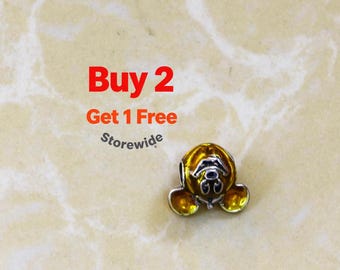 Peut inclure: Une perle breloque métallique présentant un motif de tête de souris jaune et argentée avec de grandes oreilles arrondies. Le texte Buy 2 Get 1 Free Storewide est affiché en lettres rouges et orange au-dessus de la perle sur un fond marbré clair.