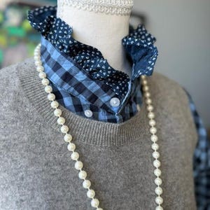 May include: A gray knit sweater layered over a blue and black gingham button-down shirt with a dark blue floral ruffled collar. A long strand of white pearls hangs over the sweater. The shirt features white buttons and a white lace trim.