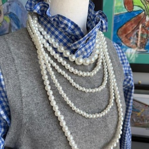 May include: A multi-strand white pearl necklace draped over a grey knit sweater vest worn on top of a blue and white gingham collared shirt with a ruffled neckline. The necklace consists of several layers of round white beads of varying sizes.