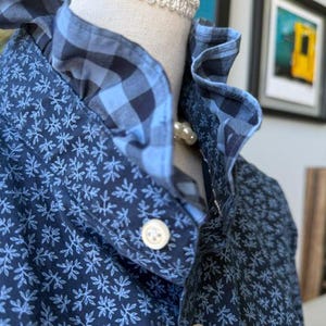 May include: A dark blue button-down shirt featuring a light blue botanical leaf pattern. The shirt has a high collar with a ruffled edge in a light and dark blue gingham check print. Two white buttons are visible on the front placket.