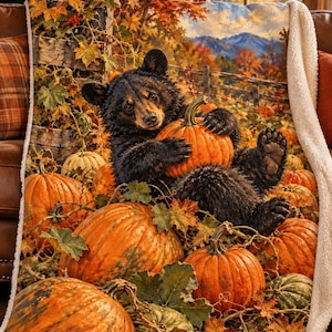 May include: A plush throw blanket featuring a printed scene of a black bear cub sitting among orange pumpkins and autumn leaves. The blanket has a white sherpa fleece lining and depicts a rustic fence and mountain landscape background.