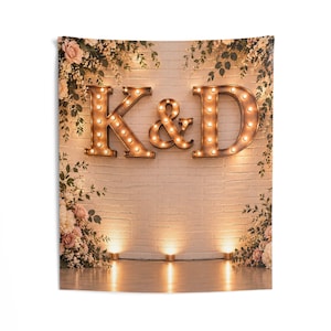 May include: A wall tapestry featuring a white brick wall backdrop with large, illuminated marquee letters K, &, and D. The display is framed by cascading floral arrangements of pink and white flowers with green leaves, accented by warm floor lighting.