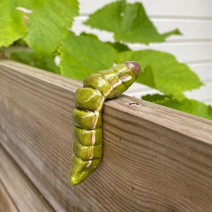 Op de afbeelding: Een glanzend, gesegmenteerd keramieken rupsbeeldje in olijfgroene tinten met witte accenten en een donkerbruine kop, gedrapeerd over een houten oppervlak. Het beeldje heeft een gladde, reflecterende afwerking en realistische segmenten.