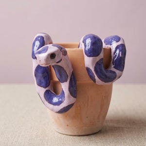 Könnte beinhalten: Ein Terrakotta-Pflanztopf mit einer geformten Keramikschlange, die sich um den Rand windet. Die Schlange ist in hellem Lavendel mit dunkelvioletten Flecken bemalt und hat eine glatte, glänzende Oberfläche. Der Topf hat eine natürliche Tontextur.