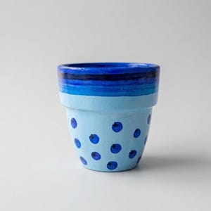 Puede incluir: Una maceta de terracota pintada a mano con una base de color azul claro decorada con puntos circulares de color azul oscuro dispersos. El borde superior de la maceta está pintado con bandas horizontales de varios tonos de azul profundo y marino.