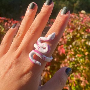 Op de afbeelding: Een ring in de vorm van een kronkelende slang, met een wit lichaam met roze vlekken en een gladde, glanzende afwerking. De slang wikkelt zich om de vinger, met de kop bij het gewricht en de staart richting de basis van de vinger.