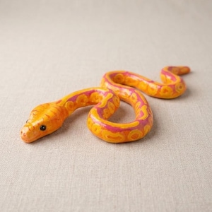 May include: A sculpted snake figurine featuring a vibrant colour palette of bright orange and hot pink. The body is covered in a repeating circular pattern, and the head displays two small, dark, glossy eyes. The snake is posed in a gentle curve.