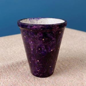 Può includere: Un vaso per piante conico con un esterno viola intenso caratterizzato da una texture maculata simile a una galassia e piccoli accenti di stelle dorate sparse. L'interno del vaso è dipinto in un bianco solido e testurizzato, e il bordo è rifinito in una tonalità viola scuro.