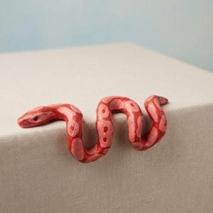 Könnte beinhalten: Eine handgeschnitzte Holzschlange mit einer leuchtend roten Grundfarbe und helleren, rötlich-rosa ovalen Flecken entlang des Körpers. Die Schlange ist in einer geschwungenen S-Form dargestellt und besitzt eine glatte, polierte Oberfläche.