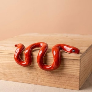 Könnte beinhalten: Eine glänzende, leuchtend rote Keramikschlange mit einer glatten, reflektierenden Oberfläche und einem einzelnen dunklen Auge. Die Schlange ist in einer gewundenen S-Form modelliert und ihr Körper liegt über der Kante einer hellen Holzkiste.