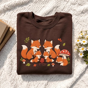 Könnte beinhalten: Ein dunkelbraunes Sweatshirt mit Rundhalsausschnitt und einer grafischen Illustration von fünf orangefarbenen Füchsen in einer Waldumgebung. Zwei erwachsene Füchse und drei kleinere Füchse sind neben einem rot-weißen Fliegenpilz und Herbstblättern abgebildet.