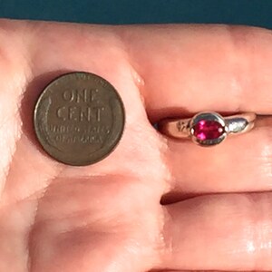 May include: A silver-toned ring featuring a small, oval-shaped, vibrant pink gemstone set in a smooth metal band, positioned next to a copper-colored United States one-cent coin for scale to demonstrate the size of the jewelry piece.