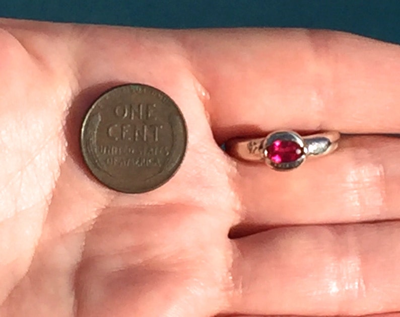 May include: A silver-toned ring featuring a small, oval-shaped, vibrant pink gemstone set in a smooth metal band, positioned next to a copper-colored United States one-cent coin for scale to demonstrate the size of the jewelry piece.