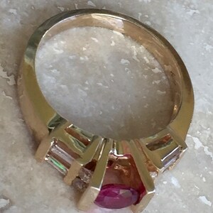 Op de afbeelding: Een gouden ring met een centrale ovaal geslepen rode edelsteen in een klauwzetting, geflankeerd door twee heldere baguette geslepen stenen aan weerszijden. De ringband is glad en gepolijst en heeft een opvallende metalen structuur.