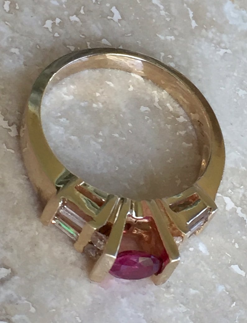 Op de afbeelding: Een gouden ring met een centrale ovaal geslepen rode edelsteen in een klauwzetting, geflankeerd door twee heldere baguette geslepen stenen aan weerszijden. De ringband is glad en gepolijst en heeft een opvallende metalen structuur.