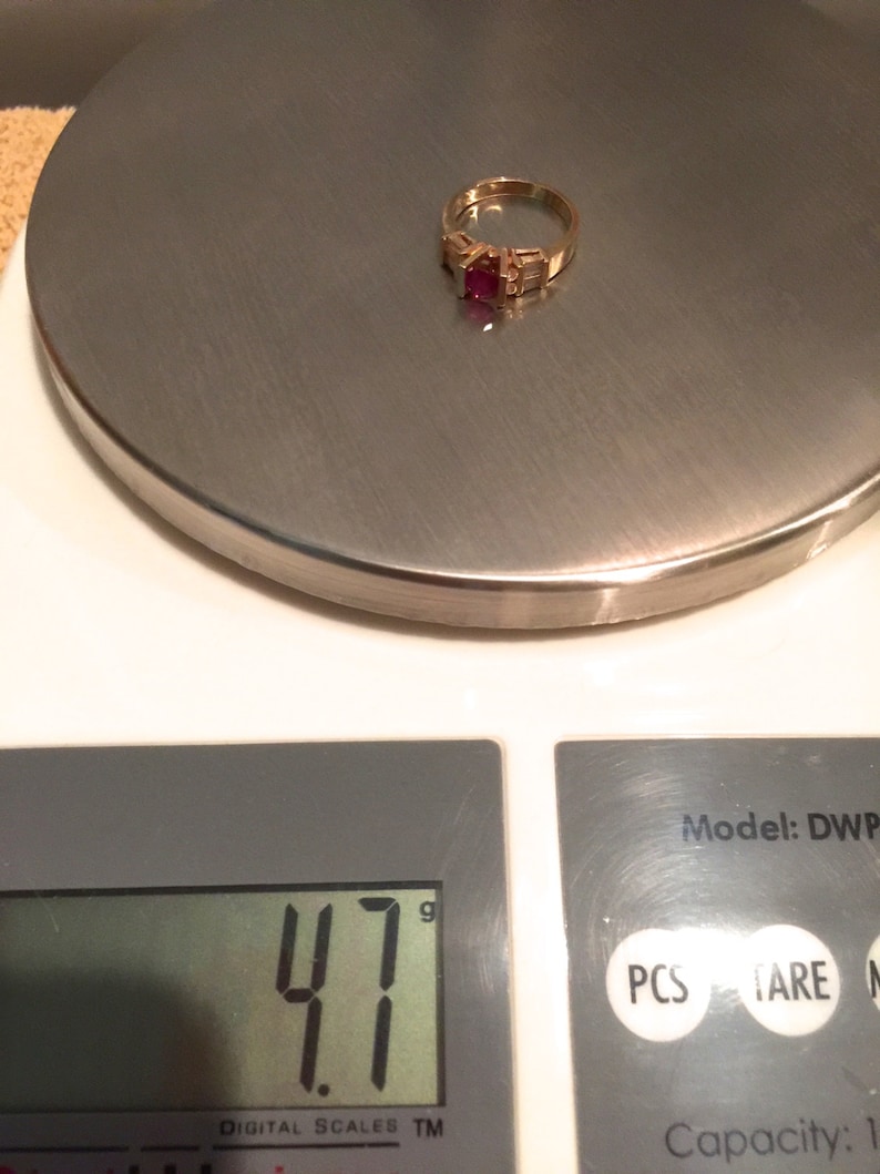 Op de afbeelding: Een gouden ring met een centrale vierkant geslepen rode edelsteen, geflankeerd door twee baguette geslepen heldere stenen aan weerszijden. De ring ligt op een digitale weegschaal die een gewicht van 4,7 gram aangeeft.