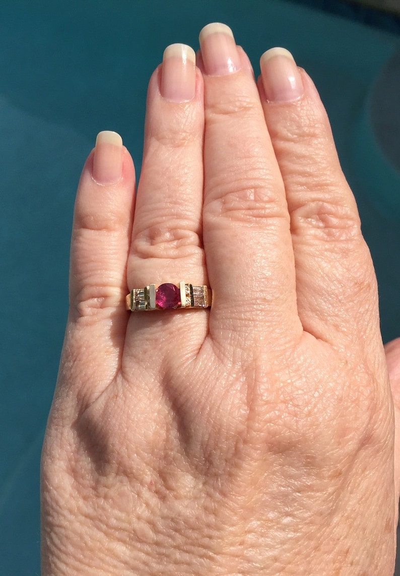 Op de afbeelding: Een gouden ring met een centrale rondgeslepen dieproze edelsteen, geflankeerd door twee rechthoekige, heldere baguette-geslepen stenen aan elke kant, gezet in een kanaalstijl band die het contrast tussen het levendige midden en de stenen benadrukt.