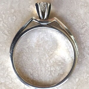 Pode incluir: Um anel de ouro com uma &uacute;nica pedra preciosa transparente de corte redondo, presa por um engaste de m&uacute;ltiplas garras. O aro &eacute; liso e circular, conectando-se ao engaste elevado no topo. O metal tem um acabamento polido e reflexivo.
