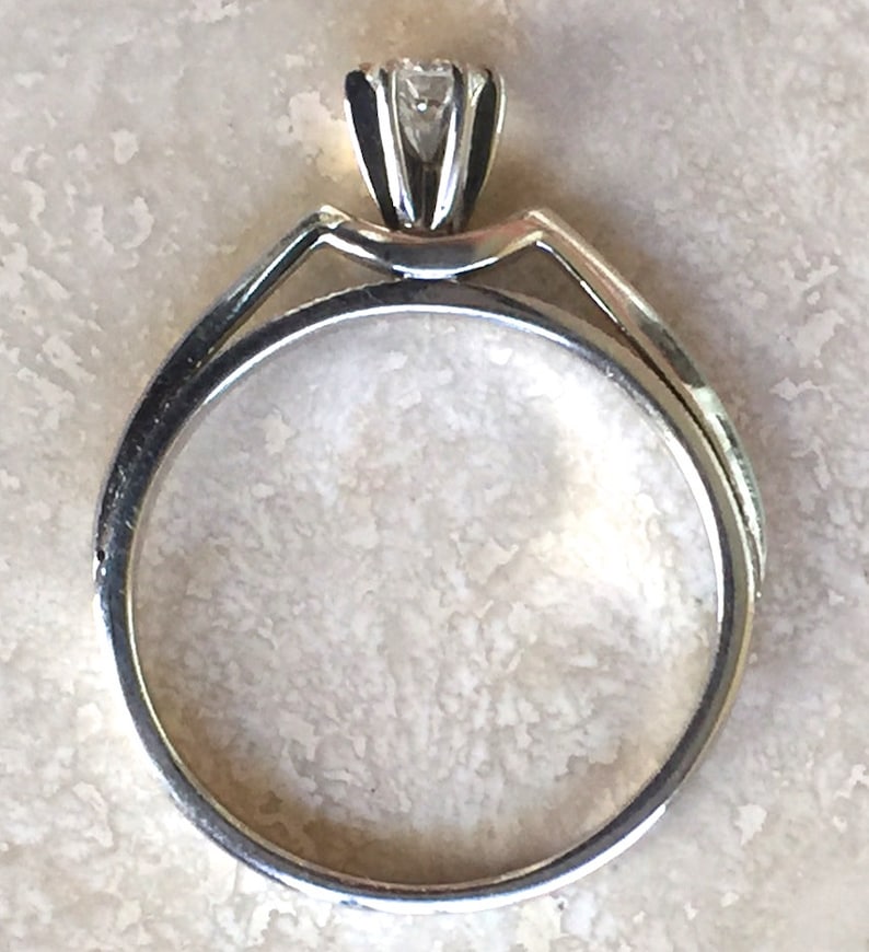 Pode incluir: Um anel de ouro com uma &uacute;nica pedra preciosa transparente de corte redondo, presa por um engaste de m&uacute;ltiplas garras. O aro &eacute; liso e circular, conectando-se ao engaste elevado no topo. O metal tem um acabamento polido e reflexivo.