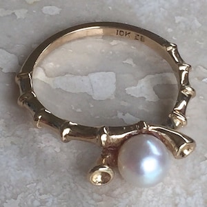 May include: A gold ring featuring a band designed to resemble bamboo segments. The ring is set with a single round white pearl and includes a small, empty cup-like setting next to it. The interior of the band is stamped with the text 10K EB.