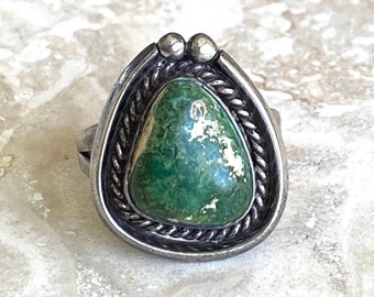 Pode incluir: Um anel de prata com uma pedra verde em forma de lágrima, apresentando padrões mosqueados em branco e verde escuro. A pedra está engastada numa borda de corda torcida e uma moldura de prata lisa, acentuada por duas pequenas esferas de prata no topo.