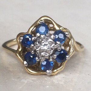 May include: A gold ring featuring a floral-shaped cluster setting. The center is a single clear stone surrounded by six round blue stones. The gold band has a wavy, openwork design that frames the central cluster of stones in a petal-like fashion.