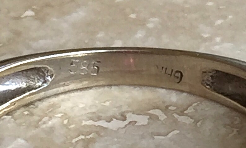 May include: A gold ring band featuring a clear, stamped hallmark on the interior surface. The metal band displays the number 585, which indicates the gold purity, along with an additional partial, partially obscured character mark next to it.