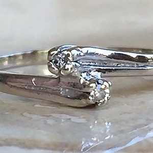 May include: A silver-toned bypass ring featuring two small, clear, round-cut stones set at the ends of the overlapping band. The metal has a polished, reflective finish, and the design creates a continuous, flowing shape across the finger.