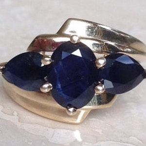 May include: A gold ring featuring a central oval-cut dark blue sapphire flanked by two pear-cut dark blue sapphires. The stones are held in place by small metal prongs, set into a polished gold band that wraps around the base of the gemstones.