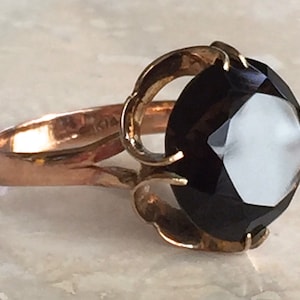 May include: A gold ring featuring a large, round, faceted dark brown gemstone held in a decorative prong setting. The band is smooth and polished, with the text 14K clearly visible on the inner surface of the metal shank of the ring.