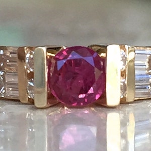 Op de afbeelding: Een gouden ring met een centrale, rond geslepen, felroze edelsteen geflankeerd door twee verticale gouden staven. Aan weerszijden van de staven zijn twee heldere baguette-geslepen stenen en twee kleine ronde heldere stenen in de band gezet.