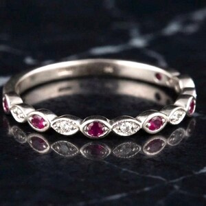 May include: A silver-toned band ring featuring an alternating pattern of round red gemstones and small clear stones set within marquise-shaped metal frames. The ring has a polished finish and a continuous design that wraps around the entire band.