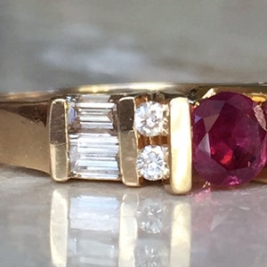Op de afbeelding: Een gouden ring met een centrale ronde rode robijn, geflankeerd door twee kleine ronde diamanten en aan weerszijden een rechthoekige diamant in baguette-slijpvorm. De stenen zijn gezet in een gouden band met een gepolijste, gladde afwerking.