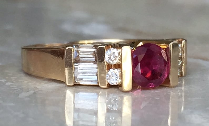 Op de afbeelding: Een gouden ring met een centrale ronde rode robijn, geflankeerd door twee kleine ronde diamanten en aan weerszijden een rechthoekige diamant in baguette-slijpvorm. De stenen zijn gezet in een gouden band met een gepolijste, gladde afwerking.
