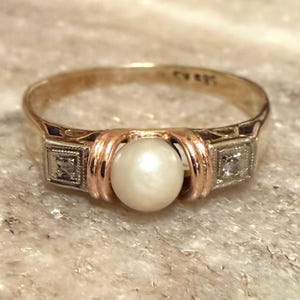 May include: A gold ring featuring a central round white pearl flanked by two small square settings, each containing a single clear gemstone. The band has a polished finish and includes a visible hallmark stamp on the inner surface.