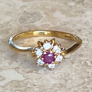May include: A gold ring featuring a central round-cut red ruby surrounded by a cluster of eight small, clear, round-cut white stones arranged in a floral pattern. The band is smooth and polished gold, with a bypass-style design leading to the setting.