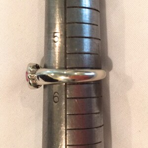 May include: A silver-toned ring with a small pink circular stone setting positioned on a metal ring mandrel. The ring rests between the 5 and 6 size markings on the tool, indicating a ring size of approximately 5.5 on the standard US scale.