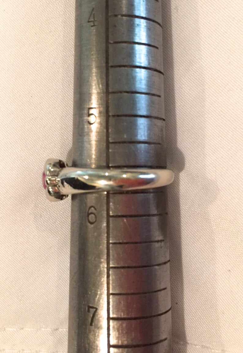 May include: A silver-toned ring with a small pink circular stone setting positioned on a metal ring mandrel. The ring rests between the 5 and 6 size markings on the tool, indicating a ring size of approximately 5.5 on the standard US scale.