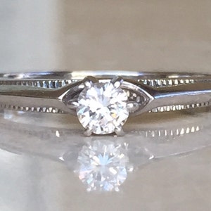 Pode incluir: Um anel de metal prateado com uma pedra redonda transparente facetada montada em quatro garras. A banda apresenta um detalhe texturizado estilo milgrain nas laterais, que leva a um design de haste lisa e c&ocirc;nica.
