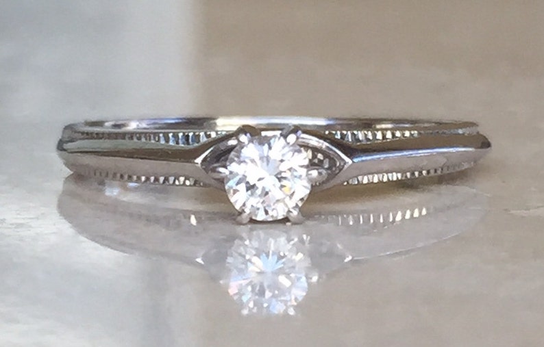Pode incluir: Um anel de metal prateado com uma pedra redonda transparente facetada montada em quatro garras. A banda apresenta um detalhe texturizado estilo milgrain nas laterais, que leva a um design de haste lisa e c&ocirc;nica.